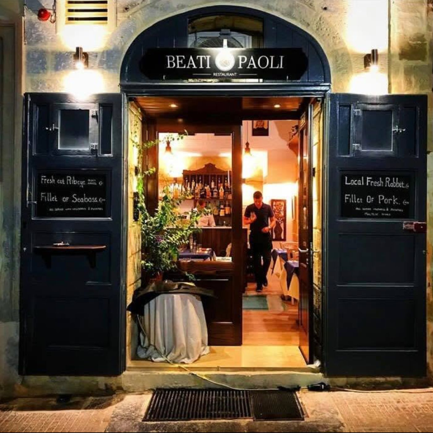 The arched entrance of Beati Paoli Restaurant at night, with open navy doors, chalkboard specials and the warmly lit dining room inside