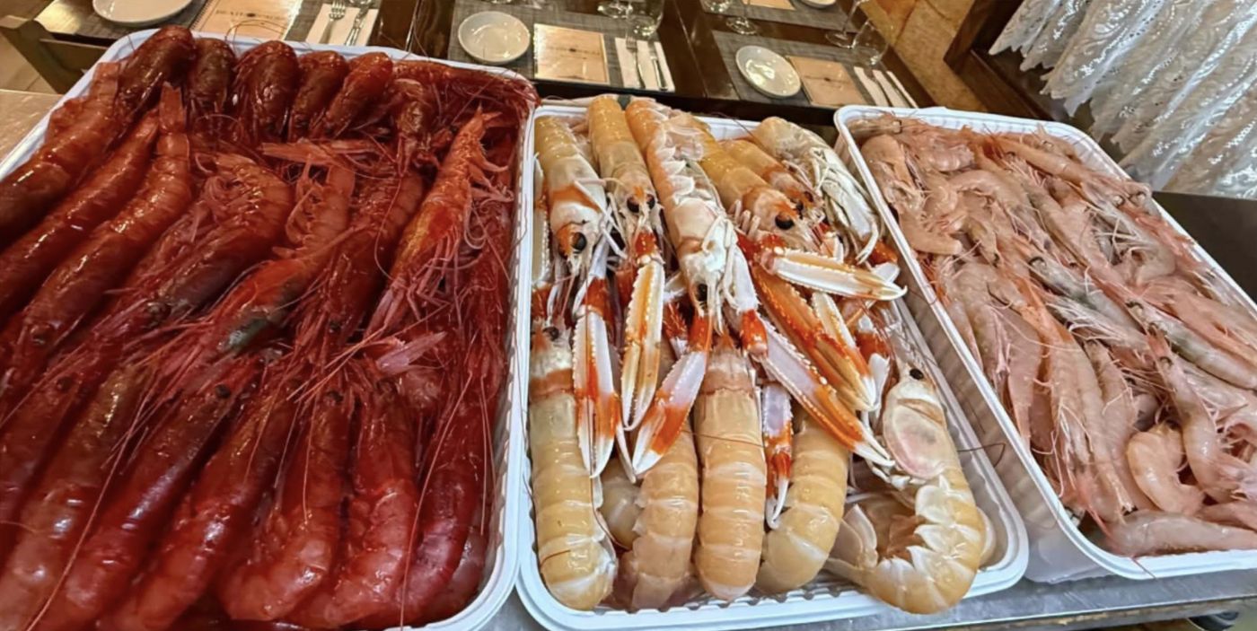 Three trays of fresh raw seafood side by side: deep red prawns, langoustines and pink prawns, with set tables behind