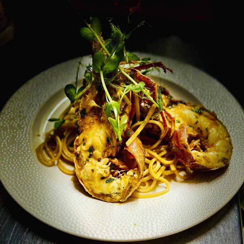 Spaghetti with a halved grilled lobster in a golden butter sauce, topped with pea shoots, on a white textured plate