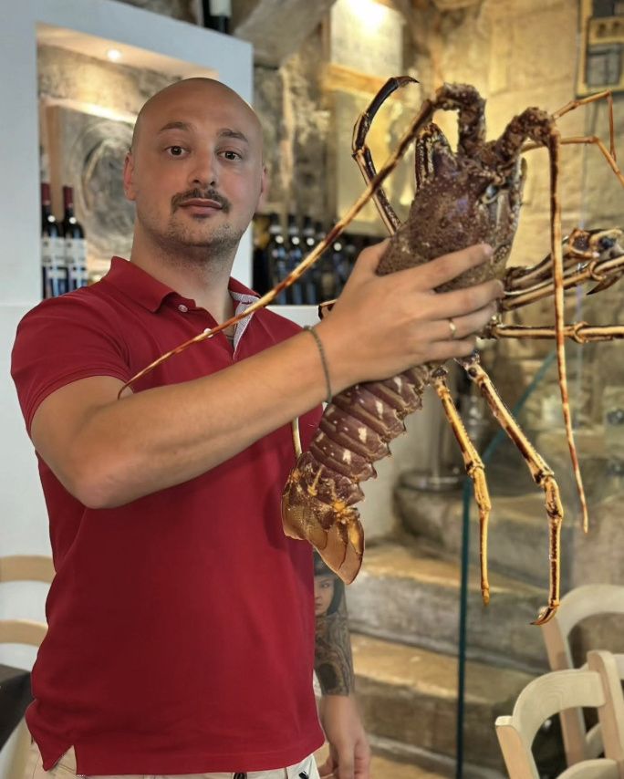 A staff member in a red polo shirt holding up a large live spiny lobster in the limestone dining room