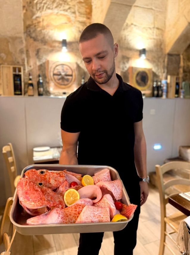 A staff member in a black shirt holding a tray of fresh red scorpionfish with lemon halves and cherry tomatoes, limestone walls behind